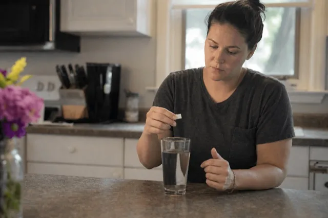 A person holding a sublingual Suboxone film over a glass of water in a calm kitchen